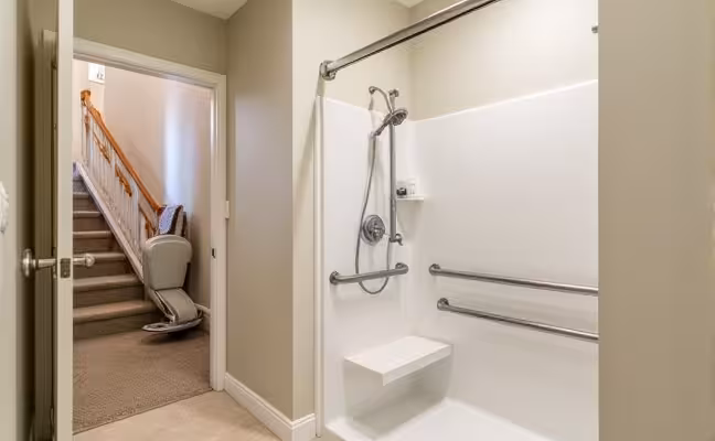 A well-lit, spacious bathroom featuring a walk-in shower with a built-in bench and grab bars. Additionally, the doorway is wide enough for wheelchair access. In the background, a stairlift is visible, installed along a staircase leading to the second floor of the home